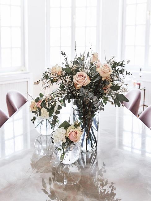 Blumendeko Tisch Heller langer Tisch in einem lichtdurchfluteten Raum, darauf drei klassische Vasen mit Rosensträußen in Pastellrosa