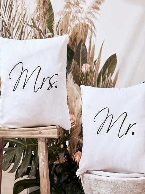 hochzeitsmotto Zwei weiße Kissen mit Mrs und Mr Aufschrift auf einem Holzhocker und einem Topf vor Pflanzendeko