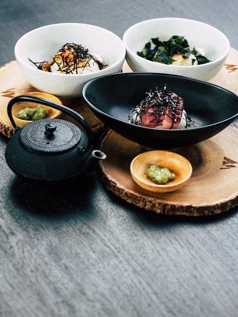 Mehrere Bowl-Schüsseln mit Essen und einer Teekanne auf Holzplatten auf dem Esstisch