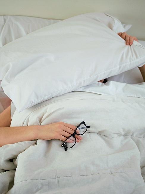 Person mit Brille in der Hand im Bett zugedeckt