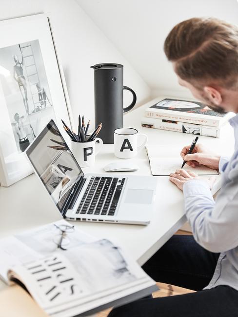 Work life balance Mężczyna siedziący przy białym biruku z laptopem, kubkiem i innymi akcesoriami do pracy