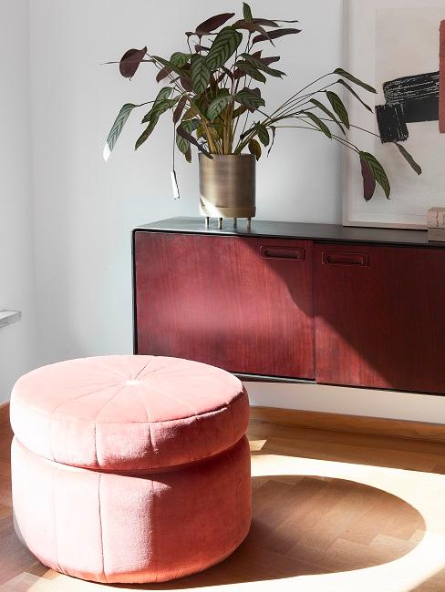 Ein Pouf steht neben einem Sideboard mit einer Toppflanze darauf in einem goldenen Topf