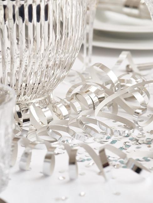 Tischdeko Silvester Gedeckter Tisch mit Luftschlangen und Konfetti in Silber auf einer weißen Tischdecke als Silvester Inspiration