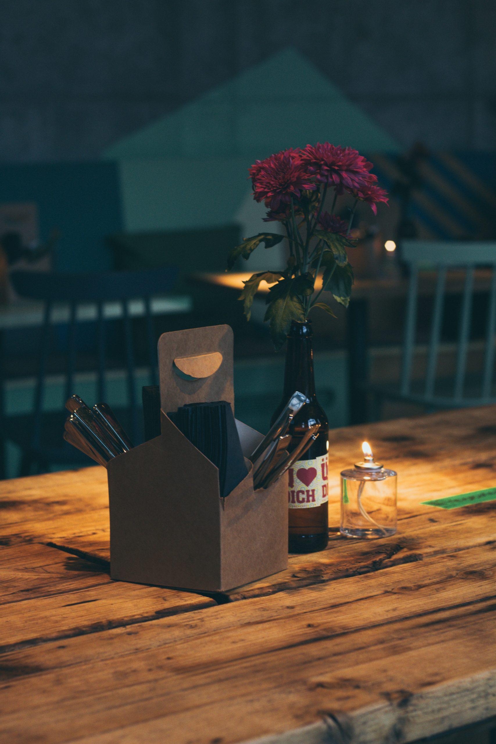 cena-romantica-banner Mesa de madera con cubiertos y flores metidas dentro de una botella de vino, junto a un portavelas de cristal con una vela encendida
