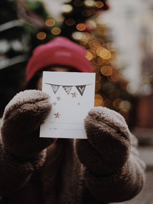 Weihnachtskarte basteln Ein Mädchen mit Mütze und Hanschuhen hält vor ihrem Gesicht eine selbstgebastelte Weihnachtskarte in die Kamera, im Hintergrund ein Weihnachtsbaum