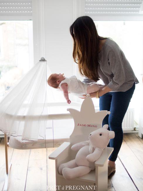 chambre mixte pour bébé une femme tenant a bout des bras un nourrisson, derriere un berceau avec baldaquin blanc