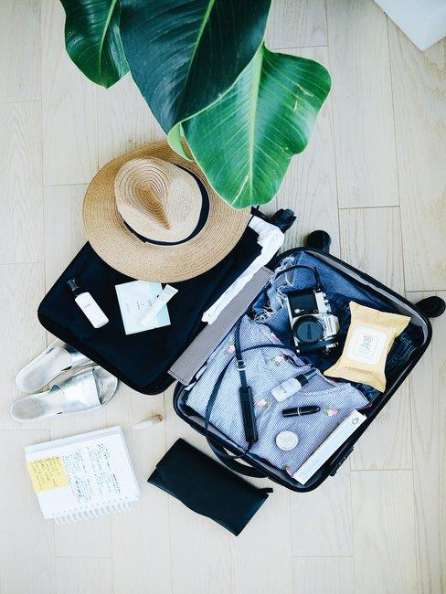 Checlist vakantie header Open koffer van boven op licht houten vloer met inhoud en rieten hoed