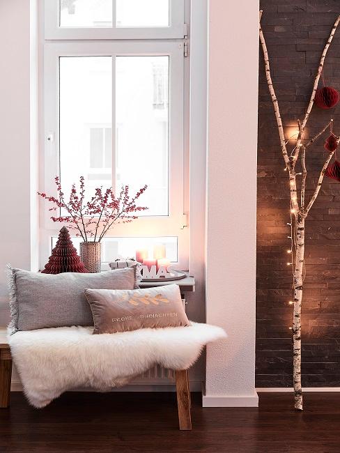 Fensterdeko Weihnachten Gemütliche Sitzbank mit Kissen und Fell vor einem Fenster mit weihnachtlicher Deko, daneben ein Baumstamm mit Lichterkette