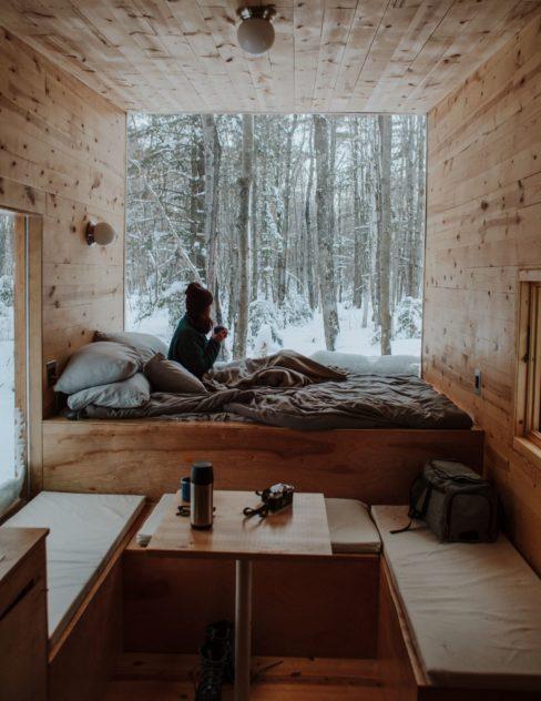 matériaux écologiques intérieur boisé d'un tiny house, une femme assise dans un lit en train de boire un café et regarder le bois enneigé à travers le grand vitre
