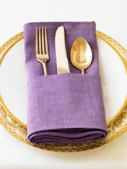 gros plan des assiettes à bord doré et couverts dorés posés dusses emballés dans une serviette de table en tissu violet
