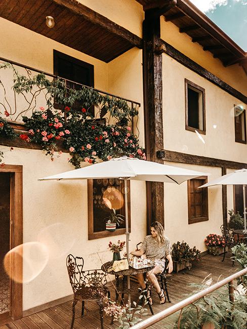 Terrasse avec personne entrain de prendre un café assise sous un parasol et un balcon fleuri au dessus