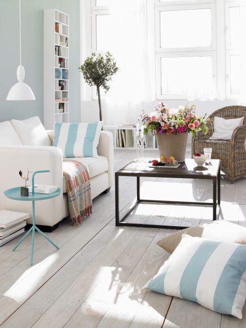 salon blanc et bleu clair, canapé blanc, coussins rayures bleues et blanches, sol en bois vieilli, table basse design avec un bouquet de fleurs pose dessus, fauteuil en rotin avec coussin blanc
