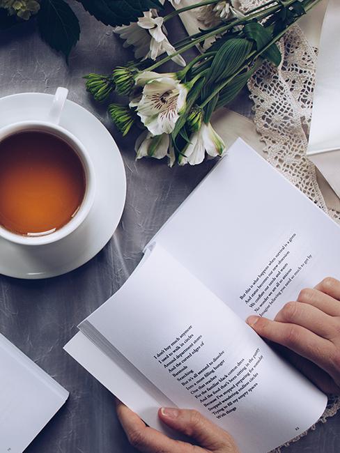 personne entrain de lire un livre avec une tasse de thé et des fleurs posées sur une table