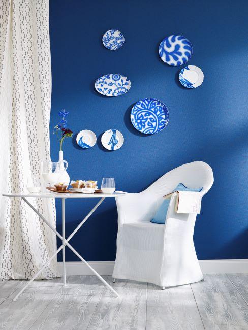 pan de mur bleu, chaise et petite table blenches, rideau blanc, décoration murale faite des plats décoratifs bleus