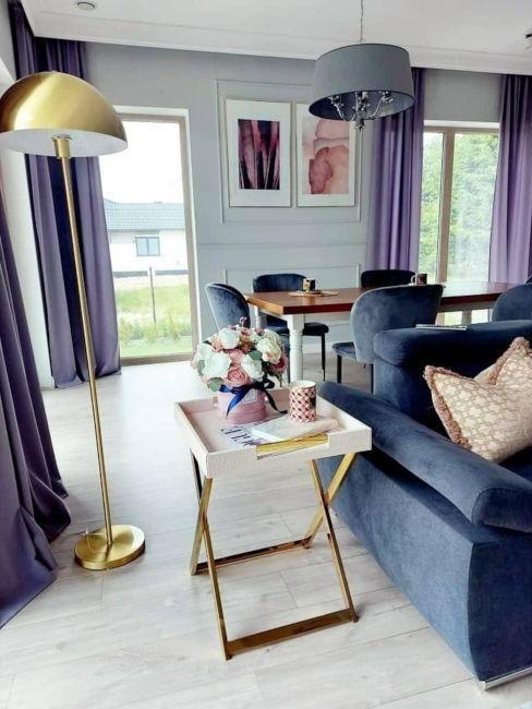 salon avec parquet clair, rideaux very peri, mur gris clair, canapé gris foncé, table à manger en bois foncé et chaise confortables gris foncé, grande lampes en métal doré et table d'appoint