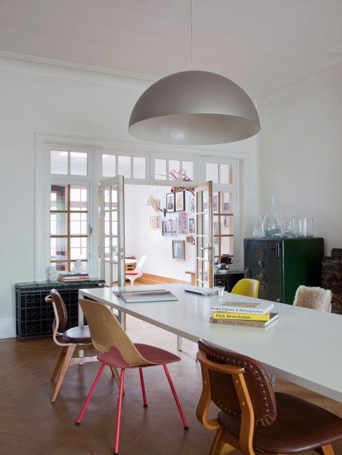 salle à manger avec grande table blanche et grande suspension grise, chaises de différentes couleurs