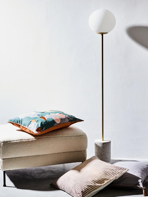 Beige voetenbank met sierkussen naast een vloerlamp van geborsteld metaal