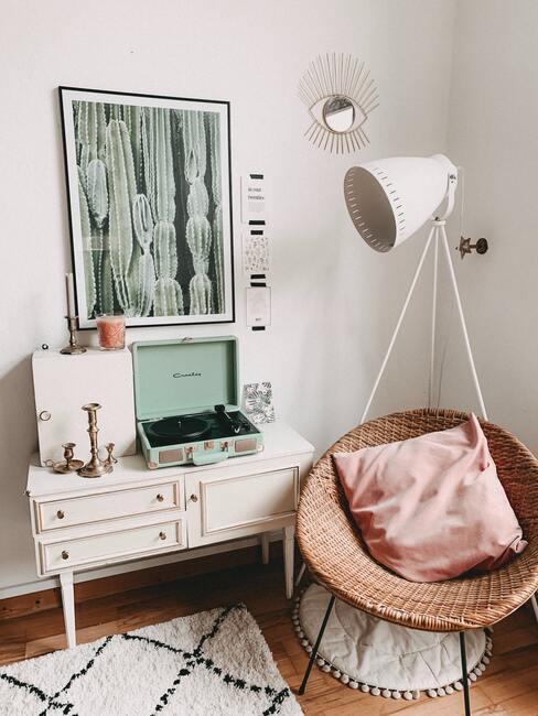 Licht vloerkleed met een geometrisch patroon naast het wit van het dressoir en de rotan stoel met een roze kussen naast een witte staande lamp