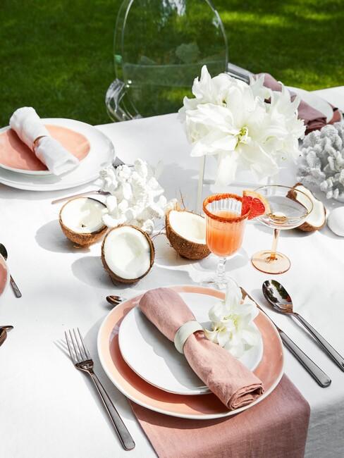 een tafel buiten met wit tafelkleed en roze, oranje en kokosnoot accessoires