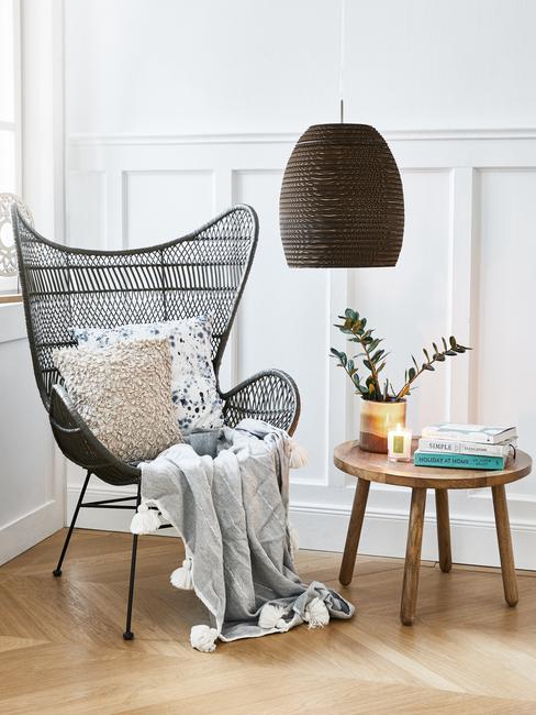 Zwarte rotan fauteuil met sierkussen en zachte plaid naast houten bijzettafel met accessoires en ornamenten