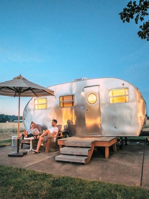 Zilveren camper van de zijkant met een stel op campingstoelen en parasol