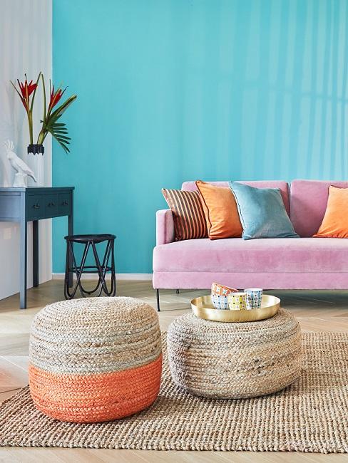 blauwe muur met een roze bank met oranje kussens en rieten met oranje poef