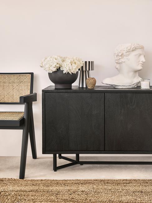 Zwarte sideboard met boeket bloemen en decoratief object naast rotan stoel