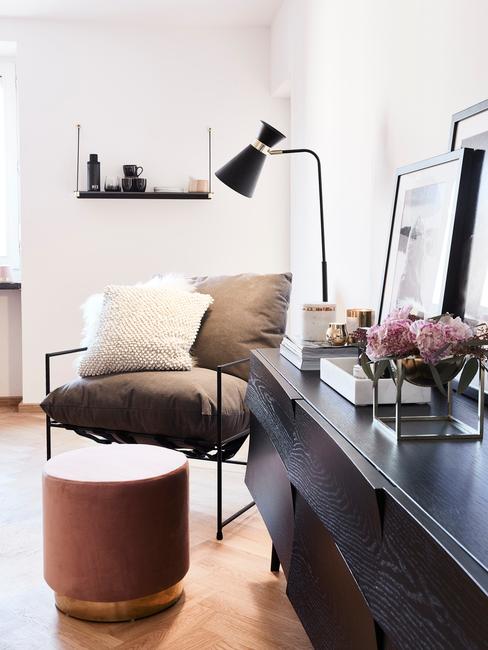 Zwarte sideboard met decoraties naast een fauteuil met sierkussens en fluwelen poef