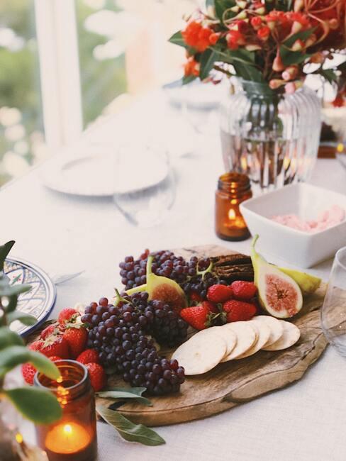 Een bord met kaas, vleeswaren en fruit op een wit tafelkleed naast een transparante vaas met bloemen