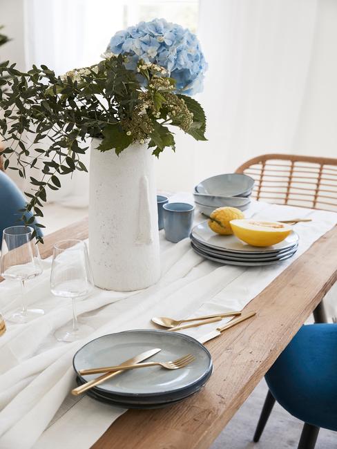 Houten eettafel met tafelkleed in wit en lichtbaluwe serviesset, witte vaas met bloemen