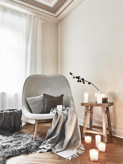 Comfortabele fauteuil in wit met sierkussen en plaid in grijs naast een houten bijzettafel met kaarsen