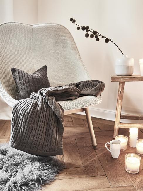 Comfortabele fauteuil in wit met sierkussen en plaid in grijs naast een houten bijzettafel met kaarsen