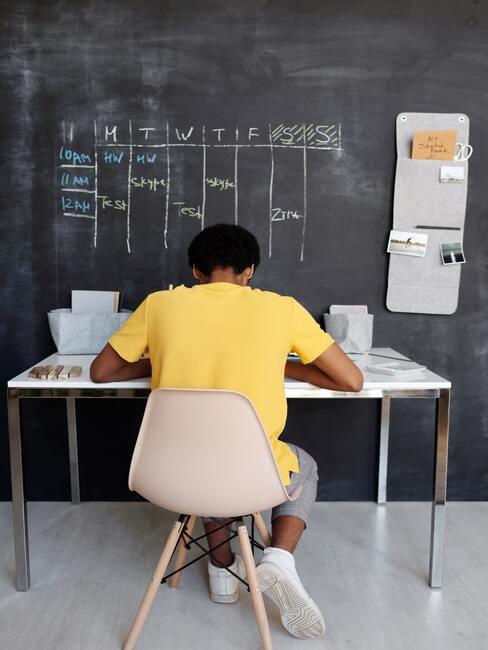 man werkt aan bureau met groot krijtbord waarop een weekschema staat