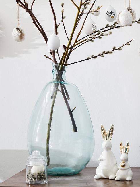 Białe figurki zajączków wielkanocnych, które stoją przy szklanym wazonie z baziami