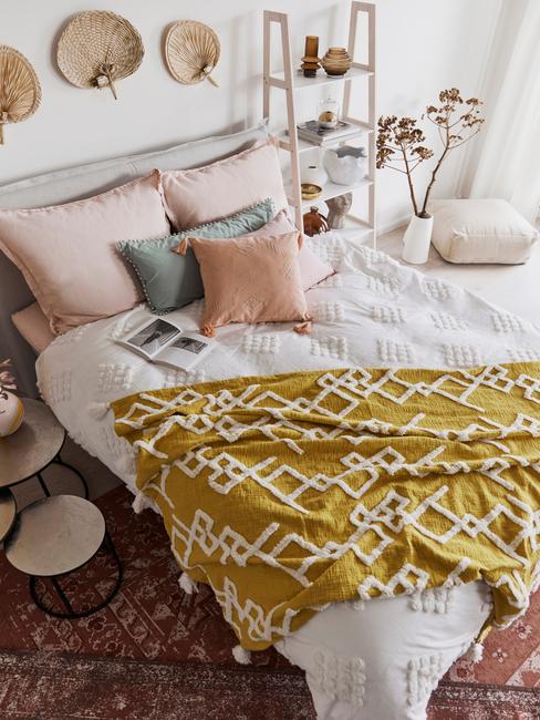 Zbliżenie na łóżko z żółtym kocem, kolorowymi poduszkami oraz słomkowym wachlarzami na ścianie