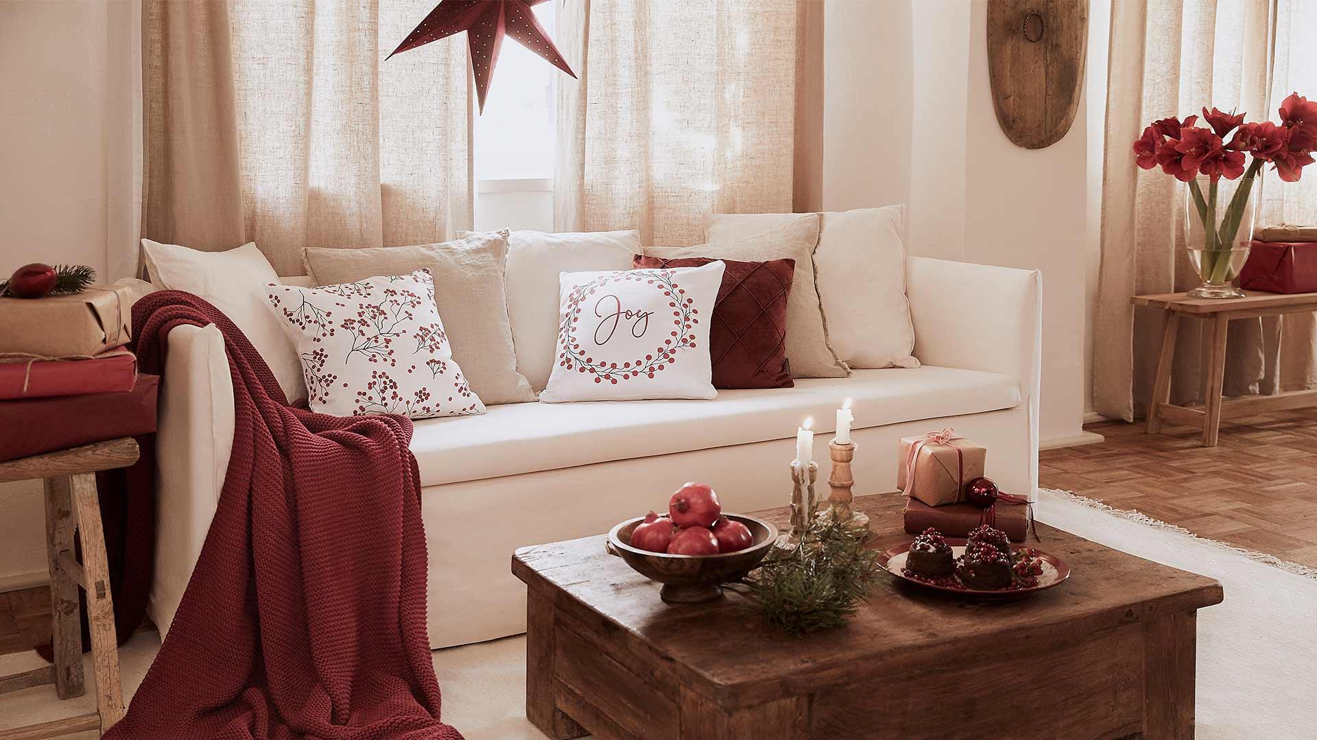 Jasny salon z beżową sofą z białymi i czerwonymi poduszkami, kocem oraz drewnianym stołem z dekoracjami