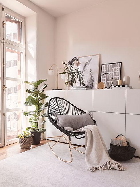 czarny bujany fotel na złotych płozach przy dużym oknie obok białej komody i dużego fikusa