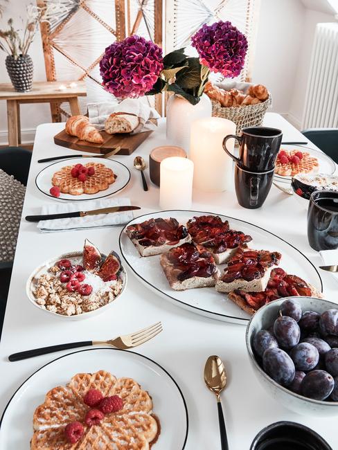 Zastawiony biały stół z zestawem białych talerzy, talerzami do serwowania oraz czarnymi kubkami na brunch