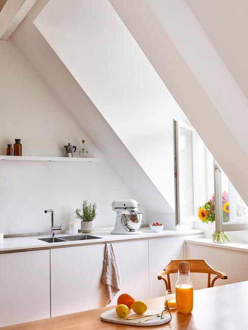 Biała kuchnia na poddaszu z białymi forntami szafek, drewnianym stołem oraz drewnianym krzesłem