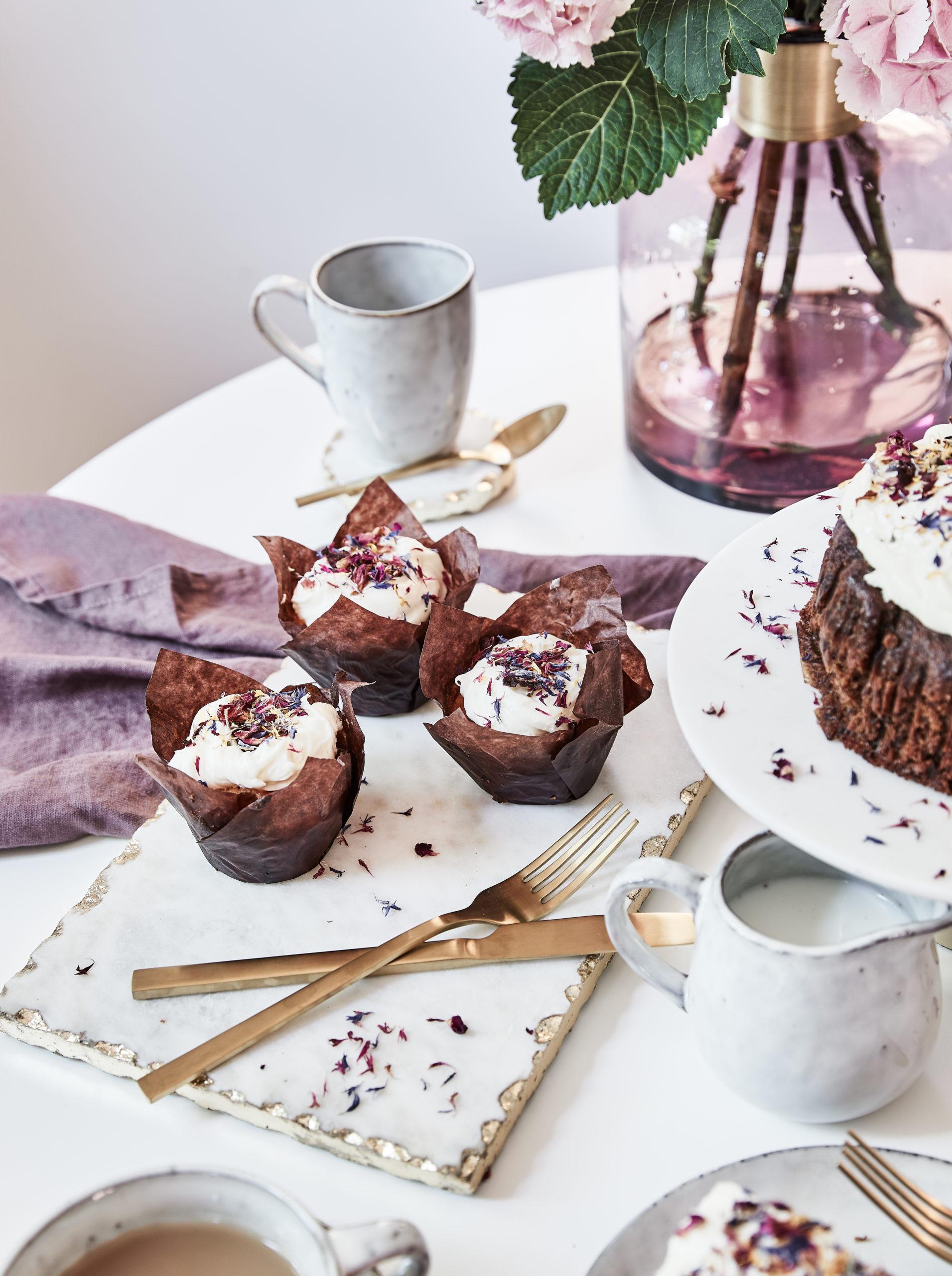 Trzy czekoladowe babeczki na stole obok białej patery z ciastem czekoladowym