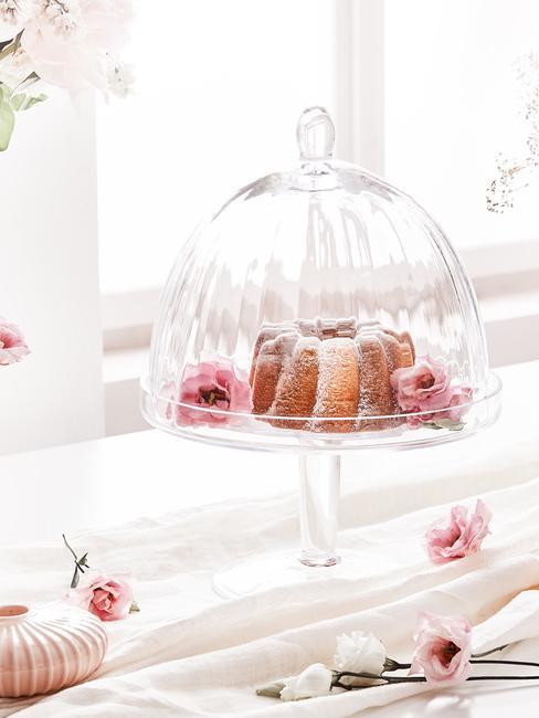 Szklana patera z ciastem oraz kwiatami ustawiona na parapecie przy oknie