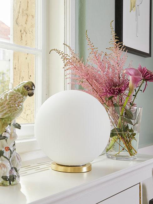 Okrągła lampa stołowa na parapecie obok porcelanowej figurki papugi obok szklanego wazonu z różowymi kwiatami
