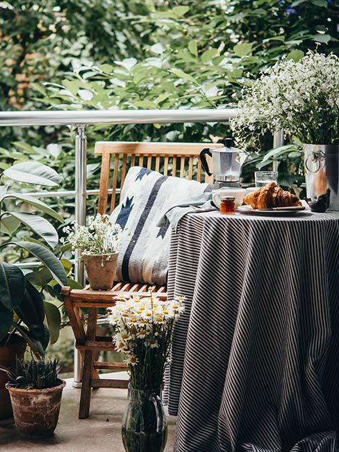 Mały balkon w zieleni z drewnianym krzesłem i nakrytym stoliczkiem z kawiarką i rogalikiem