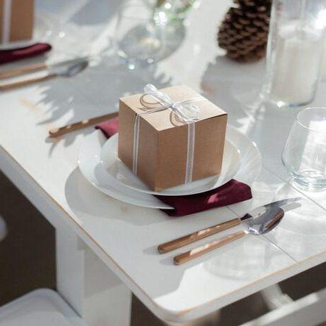 Biała zastawa stołowa na stole, na której położne jest małe małe złote pudełko, które jest prezentem dla gości