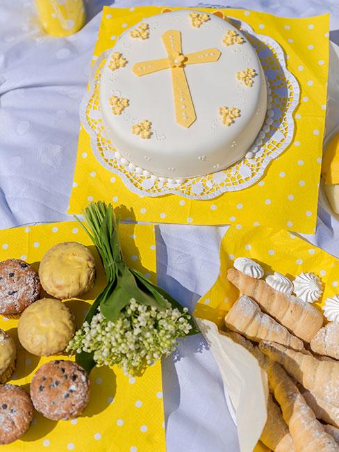 jasnożółty tort na chrzest na żółtmy obrusie obok pączków i bukiecika konwalii