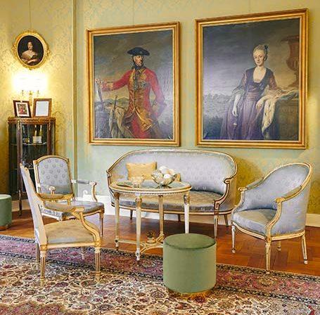 Salon z wzorzystym dywanem, zestawem mebli ze złotym rantem i błękitnym obiciem i dwoma obrazami na ścianie w złotych ramach