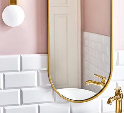 Różowa ściana w łazience, białe płytki i złote dodatki w postaci lampy i lustra