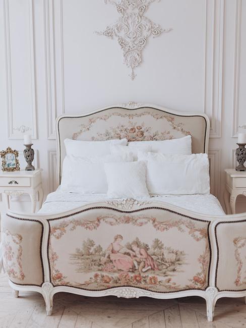 Bogato zdobione lóżko z malunkami z przodu i na wezgłowiu, biała pościel, białe stoliki nocne ze świecznikami