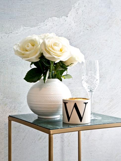 Biały wazon na stoliku ze złotymi nogami, obok świeczka w białym opakowaniu z literą"w"