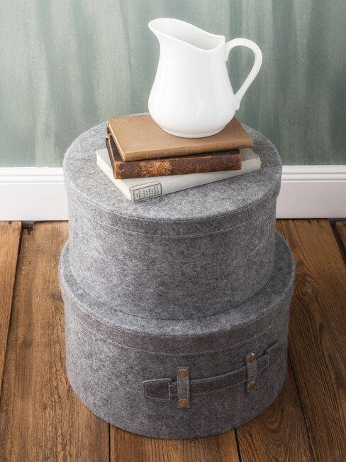 Dwa pudełka w szarym kolorze na których leżą książki i biały dzbanek